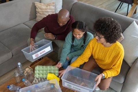 Multiracial family sorting recyclables on sofa using labeled glass and paper bins