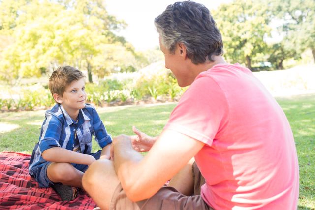 Father and son enjoying a sunny day at the park, sitting on a picnic blanket and having a meaningful conversation. Ideal for use in family-oriented advertisements, parenting blogs, and articles about spending quality time with children.