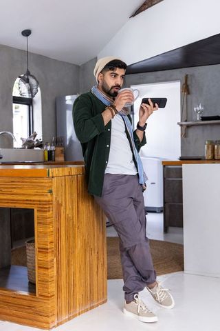 Indian man leaning on wooden kitchen island sipping coffee and checking smartphone