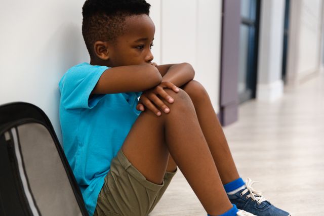 Sad african american elementary schoolboy looking away while sitting on floor in corridor. unaltered, childhood, education, sadness, loneliness, failure and school concept.