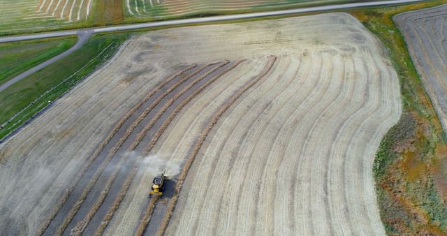 An impressive aerial view showing a harvester actively working on a large, golden agricultural field. The orderly rows of harvested crops and clear patterns in the earth provide a sense of structured farming practices. This image is excellent for illustrating modern farming machinery, agricultural activities, and rural landscapes. Perfect for usage in agricultural reports, farming lifestyle blogs, agribusiness advertisements, and educational materials on modern farming techniques.