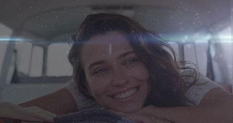 Smiling woman relaxing in camper van leaning on folded blanket with sun flare and cozy vibes