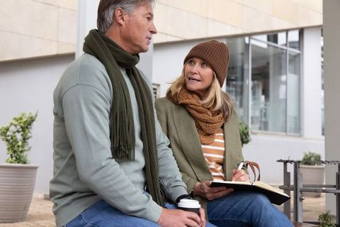 Mature couple chatting on urban bench, sharing coffee and journaling in cozy autumn attire