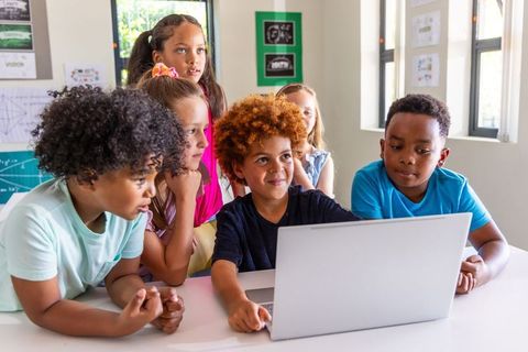 Multicultural kids collaborating around laptop in bright classroom learning together