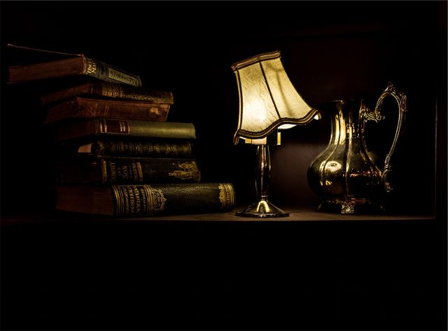 This photo captures a cozy, dimly lit scene featuring a stack of vintage books, a decorative lamp, and an ornate silver jug. It conveys a sense of nostalgia and timeless elegance, perfect for use in designs focused on literature, history, and classic home decor. Ideal for blog posts, social media, or website banners relating to reading, antiquarian aesthetics, or the cozy ambience of a study.