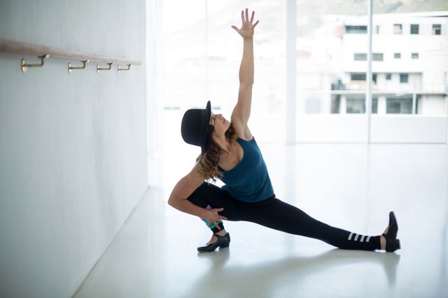 Woman dancer practicing a stretching routine in a bright dance studio. She is wearing a hat, tank top, and leggings, showcasing flexibility and balance. Ideal for use in articles or advertisements related to dance, fitness, healthy lifestyle, and training programs.