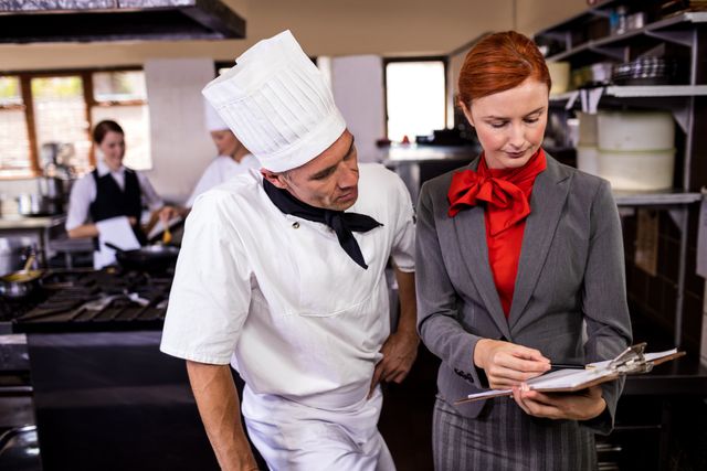 Female manager and male chef discussing and writing on clipboard in hotel kitchen. Ideal for illustrating teamwork, hospitality industry, restaurant management, and professional collaboration in culinary settings.