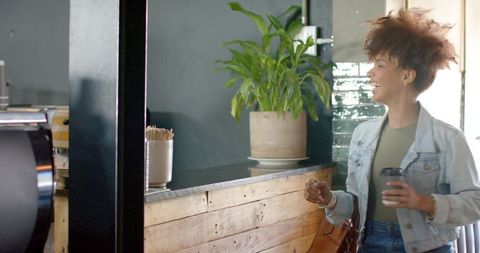 African american woman laughing at cafe counter holding takeaway coffee cup in denim jacket