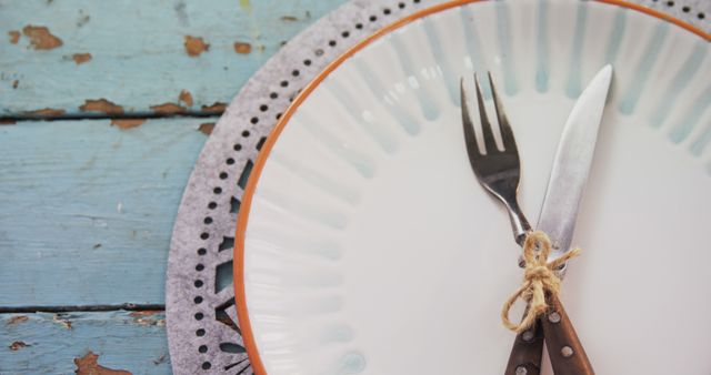 A rustic table setting features a white plate with a decorative border, accompanied by a fork and knife tied together with twine, with copy space. The vintage charm of the setting suggests a cozy, homey dining experience or a country-style event.