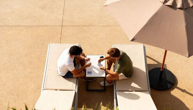 Two friends playing cards on sunlit pool terrace lounge chairs under umbrella