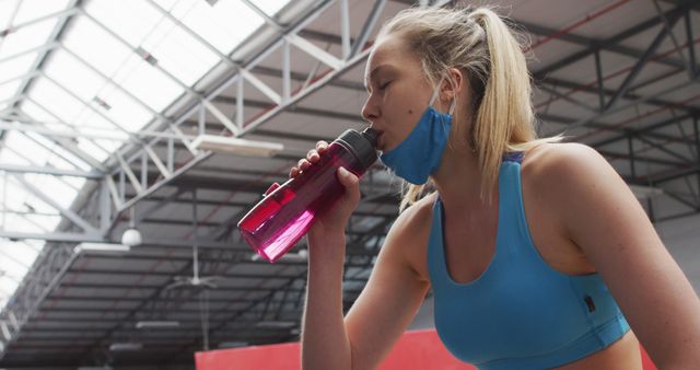 Caucasian woman wearing lowered face mask drinking water at gym. blonde woman taking a break from her workout, drinking from water bottle. hygiene at gym during coronavirus covid 19 pandemic