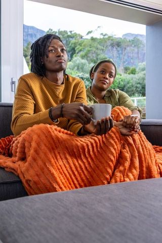 African american couple cuddling under orange throw on sofa holding mug watching movie