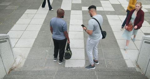 Two men standing on city plaza steps checking smartphone while holding olive duffel bag