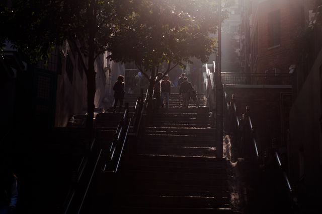 Group of people walking up stairs in an urban alleyway, with sunlight streaming through trees. This can be used for concepts related to travel, everyday city life, the urban environment, commuting, or illustrating the journey and effort themes.