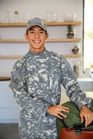Smiling asian soldier in digital camouflage uniform standing in kitchen with duffel bag