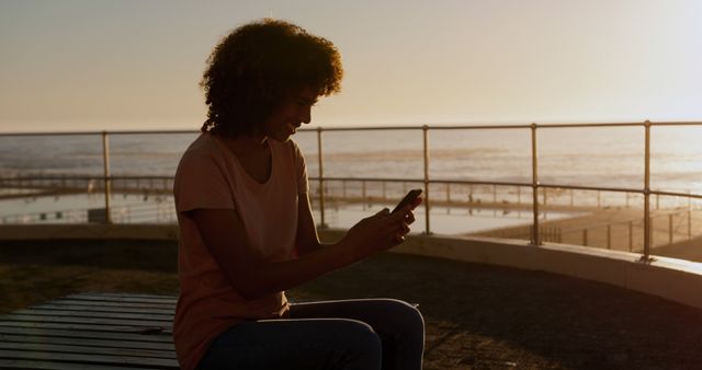 Young woman sitting on bench near the ocean at sunset, using a smartphone. Suitable for themes like relaxation, technology, communication, summer activities, outdoor leisure, travel, and connecting with nature.