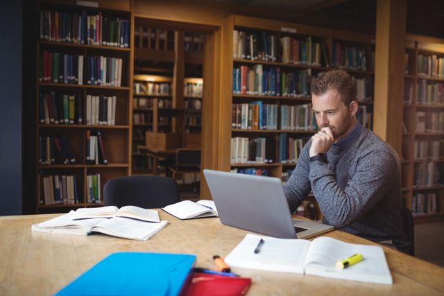 Mature student deeply focused on studying with a laptop in a college library. Surrounded by open books and study materials, he is engaged in research and learning. Ideal for use in educational content, adult education promotions, academic resources, and articles on lifelong learning.