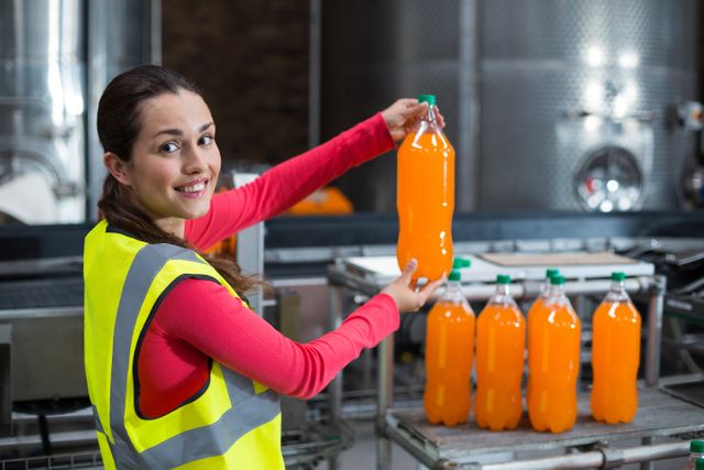 Female factory worker holding a bottle of juice in a beverage plant. She is wearing a safety vest and smiling, indicating a positive work environment. Ideal for use in articles or advertisements related to manufacturing, food and beverage industry, quality control, and industrial work environments.