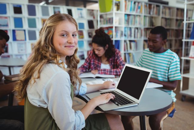 Schoolgirl smiling while using laptop in a library with classmates studying in the background. Ideal for educational content, school promotions, technology in education, group study sessions, and academic research themes.