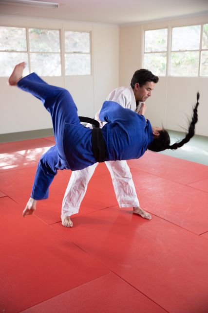 Caucasian male and female judokas wearing blue and white judogi, practicing judo on a mat during a training in a bright studio.