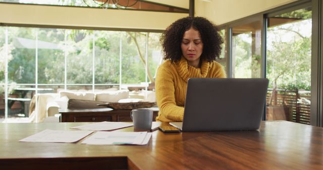 Woman working on laptop from home, seated at dining table. Modern living room in background with large windows and lots of natural light. Can be used for themes of remote work, home office productivity, and lifestyle.