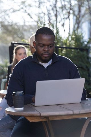 African american man working on laptop remotely at sunlit outdoor cafe with coffee cup