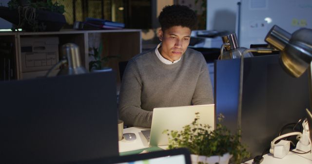 Image of focused biracial man using laptop, working late in office. Business and working in office at night with technology concept.