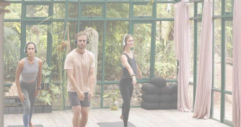 Trio practicing yoga with headphones in sunlit glass studio surrounded by plants