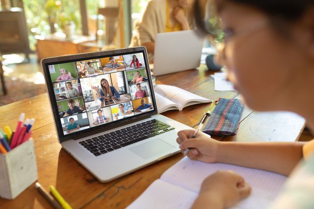 Teenage Asian girl engaging in an online class from home, laptop screen showing multiple participants on a video call. She is writing in a notebook and focusing on her studies, making use of wireless technology for remote learning. Ideal for education and e-learning websites, articles on remote education, or promotional materials for online learning platforms.