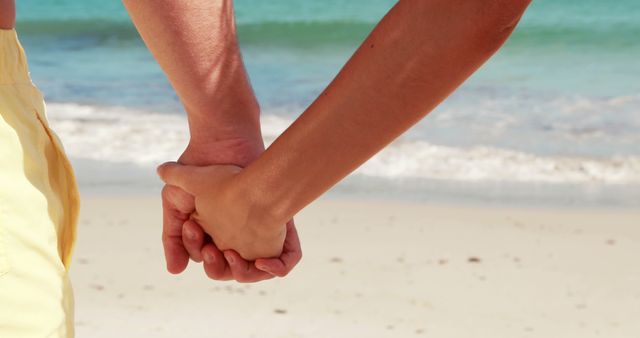 A close-up view of a couple holding hands on a beach, with copy space. Their interlocked fingers symbolize connection and affection against the backdrop of the ocean.