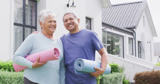 Elderly couple holding yoga mats and smiling outside their modern house. Ideal for illustrating senior wellness, healthy lifestyle choices, exercise in retirement, and activity among older adults. Suitable for advertisements, health campaigns, and magazines focusing on senior living or fitness.