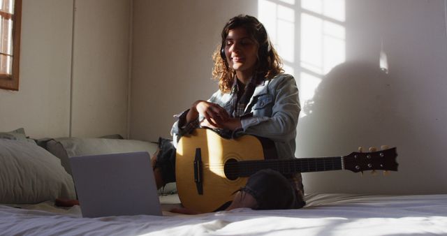 Young woman in casual clothing relaxing on bed with an acoustic guitar and using a laptop, enjoying music through headphones. Sunlight streaming through the window creates a warm, cozy atmosphere. Perfect for topics related to home leisure, music practice, relaxation, and comfortable living.