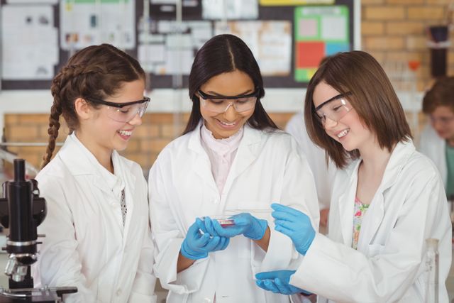 Three schoolgirls wearing lab coats and safety goggles are conducting a chemical experiment in a school laboratory. They are smiling and engaged, showcasing teamwork and collaboration in a STEM education setting. This image is ideal for educational materials, science-related articles, and promoting STEM programs for young students.