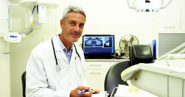 A middle-aged Caucasian male dentist is smiling at the camera in a dental clinic, with copy space. He is wearing a white lab coat and holding dental tools, with an X-ray image displayed on the monitor behind him.