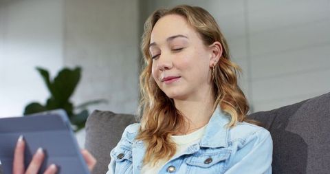 Young woman relaxing on sofa holding tablet folio in modern cozy living room, calm lifestyle