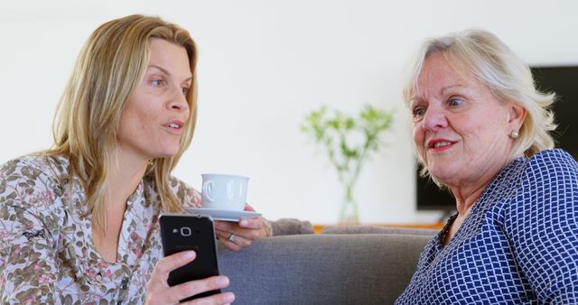 Middle-aged woman and senior woman having a conversation over coffee and using a mobile phone. Perfect for themes related to family, bonding, informal discussions, casual settings, and enjoying leisure time at home.