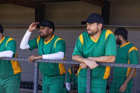 Baseball players focused watching game from dugout, leaning on railing in green uniforms