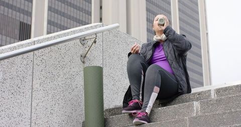 Senior woman sitting on urban steps drinking water, wearing headphones, foam roller nearby