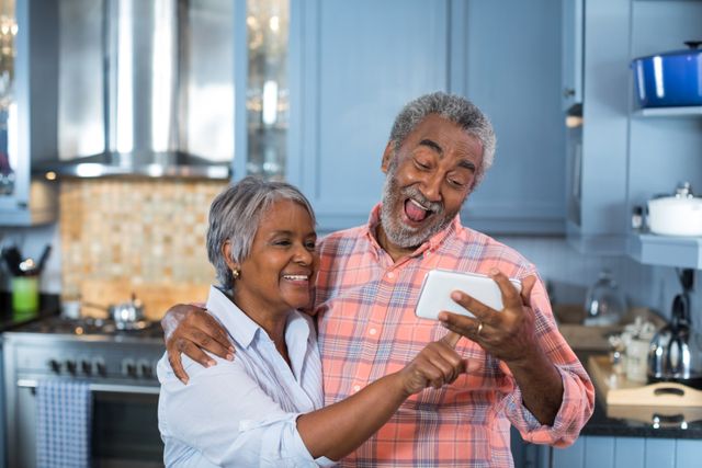 Senior couple standing in a modern kitchen, taking a selfie, and smiling. This image can be used for lifestyle blogs, articles focusing on elderly joy and technology, or advertisements promoting senior living communities.