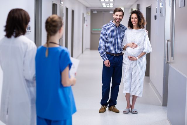 Expectant couple smiling and interacting with medical staff in a hospital corridor. The woman is wearing a hospital gown, indicating she is a patient, while the man stands supportively beside her. This image can be used for healthcare, maternity, family support, and prenatal care themes.
