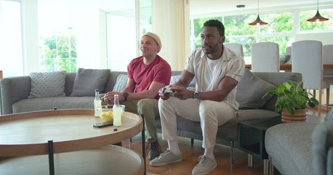 Multiracial friends playing video games on sofa in bright living room holding controllers