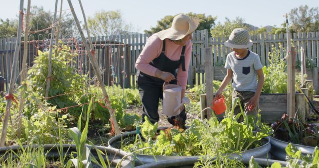 Mother and son are gardening together in a vibrant community garden. Both are wearing straw hats and are seen watering plants amidst lush green foliage on a sunny day. This image is ideal for promoting family bonding activities, sustainable living, and community engagement. It can also be used in educational materials about gardening and organic farming.