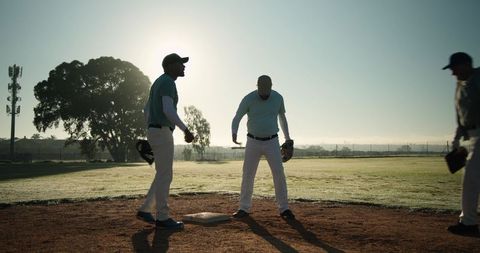 Silhouetted Baseball Players and Coach on Dewy Infield at Dawn Practicing Near Base