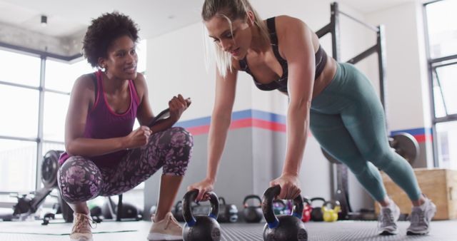 Female personal trainer coaching woman doing kettlebell push-ups at modern gym. Ideal for use in articles, blogs, and advertisements related to fitness coaching, strength training programs, personal training services, and the benefits of regular exercise.