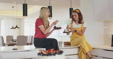 Two friends sharing berry cake on modern kitchen island, casual outfits, sunny open-plan