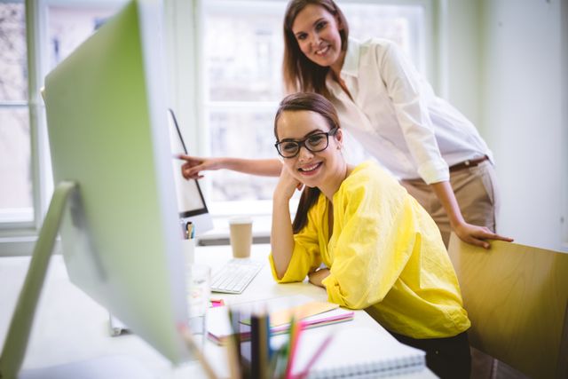 Two female colleagues are collaborating in a modern, creative office environment. One is sitting at a desk with a computer, smiling at the camera, while the other stands beside her, pointing at the screen. Both are dressed in casual attire, creating a relaxed yet professional atmosphere. This image is ideal for illustrating teamwork, collaboration, and a positive work environment in business presentations, websites, and promotional materials.