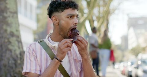 Young indian man enjoying chocolate pastry on city sidewalk with headphones and bag
