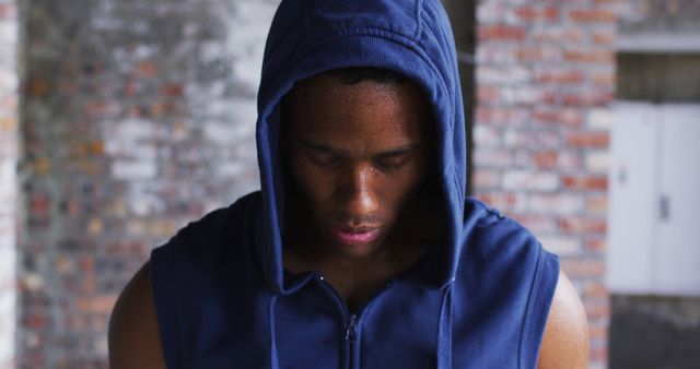 An athletic man in a blue hoodie, standing indoors with head down, in a focused and determined state of mind. This image is perfect for use in promotional materials for fitness programs, gym advertisements, motivational posters, sportswear catalogs, and websites related to physical fitness and personal training.