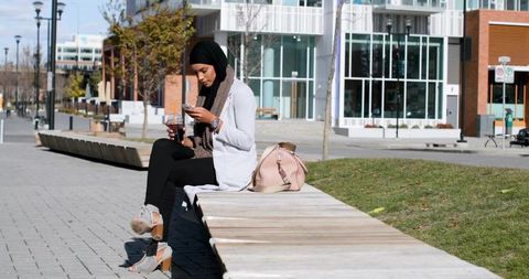 Young woman wearing hijab sitting on urban bench checking smartphone holding takeaway cup
