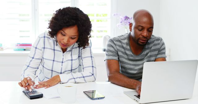 African American woman and man are focused on their tasks, with the woman using a calculator and the man working on a laptop. They appear to be professionals managing finances or working from a home office.